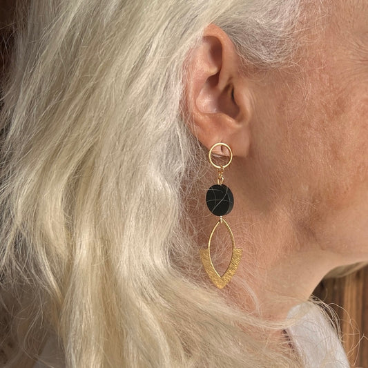 Photo of a white haired woman from the side wearing Handcrafted gold and black earrings. An open circle finding leads to a single black bead then a hammered brass marquise. 