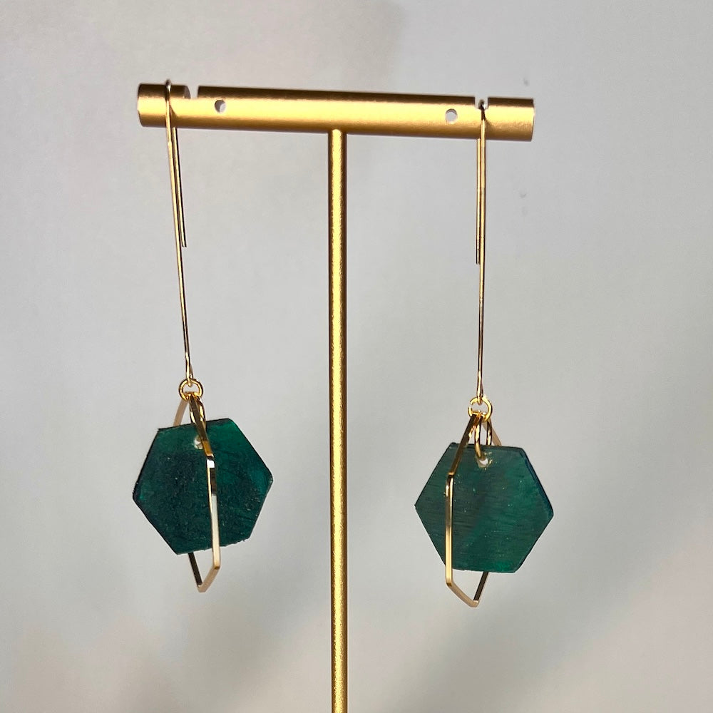 Hand painted wild seas blue- green hexagon wood bead hangs from a rigid gold threader earring - hanging from an earring display. Handcrafted by Madera Design Studio 