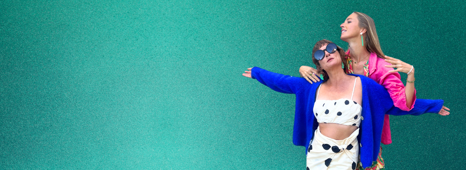 Two women posing against a teal background wearing bold fun colorful outfits and mad era jewelry