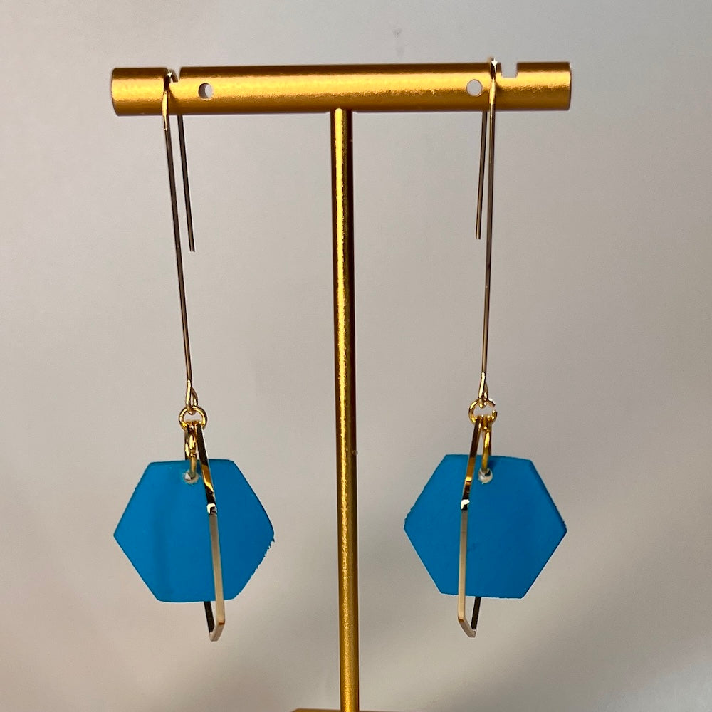 Blue hexagonal earrings on a gold earring stand against a gray background