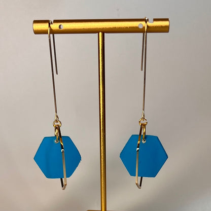 Blue hexagonal earrings on a gold earring stand against a gray background