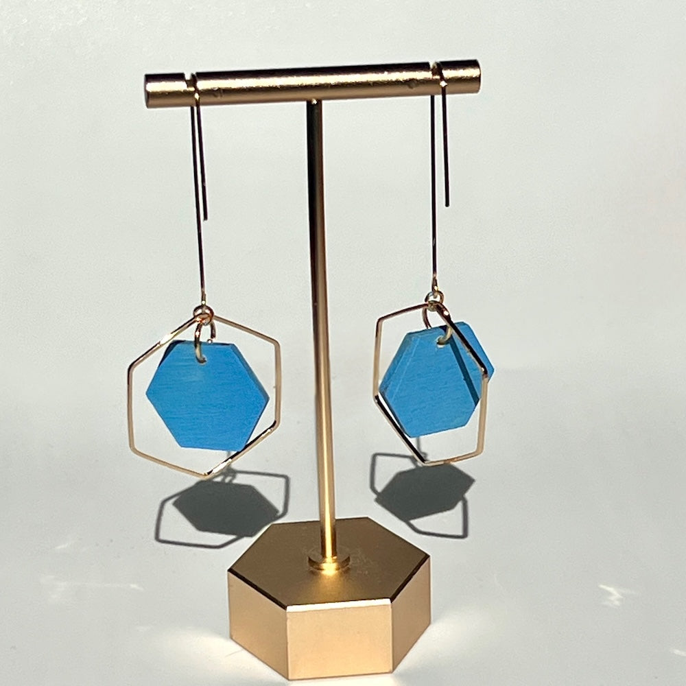 Sky Blue hand painted hexagon earrings on a white background, hanging from a golden display stand. Handcrafted by Madera Design Studio