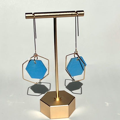 Sky Blue hand painted hexagon earrings on a white background, hanging from a golden display stand. Handcrafted by Madera Design Studio