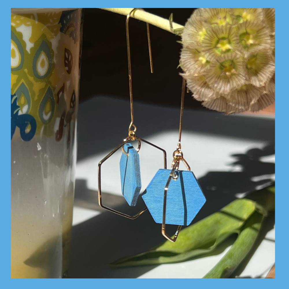 Sky Blue handpainted wooden hexagon beads hang inside a gold plated hexagon , they dangle from a rigid threader earring, and are hanging from a flower in this photo. Handcrafted by Madera Design Studio
