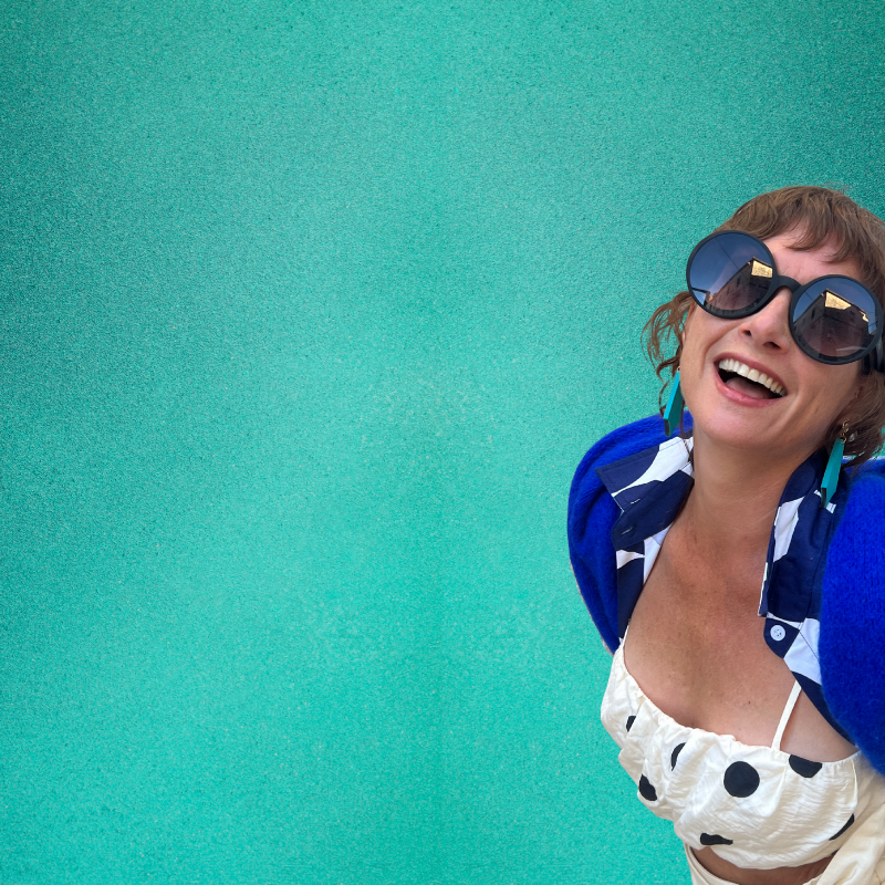 Woman wearing sunglasses a polka dot top and Mad Era painted earrings against a teal background