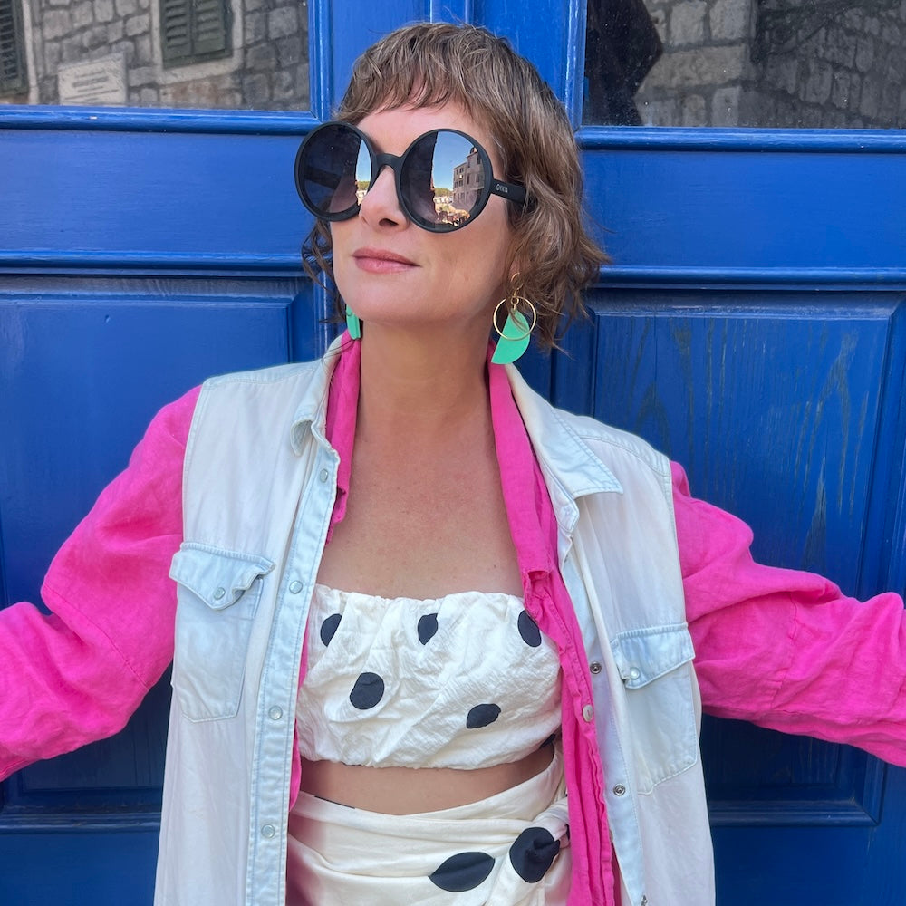 model in bright pink shirt, jean vest and polka dot bandeau wearing mad era green half tone earrings on a blue door