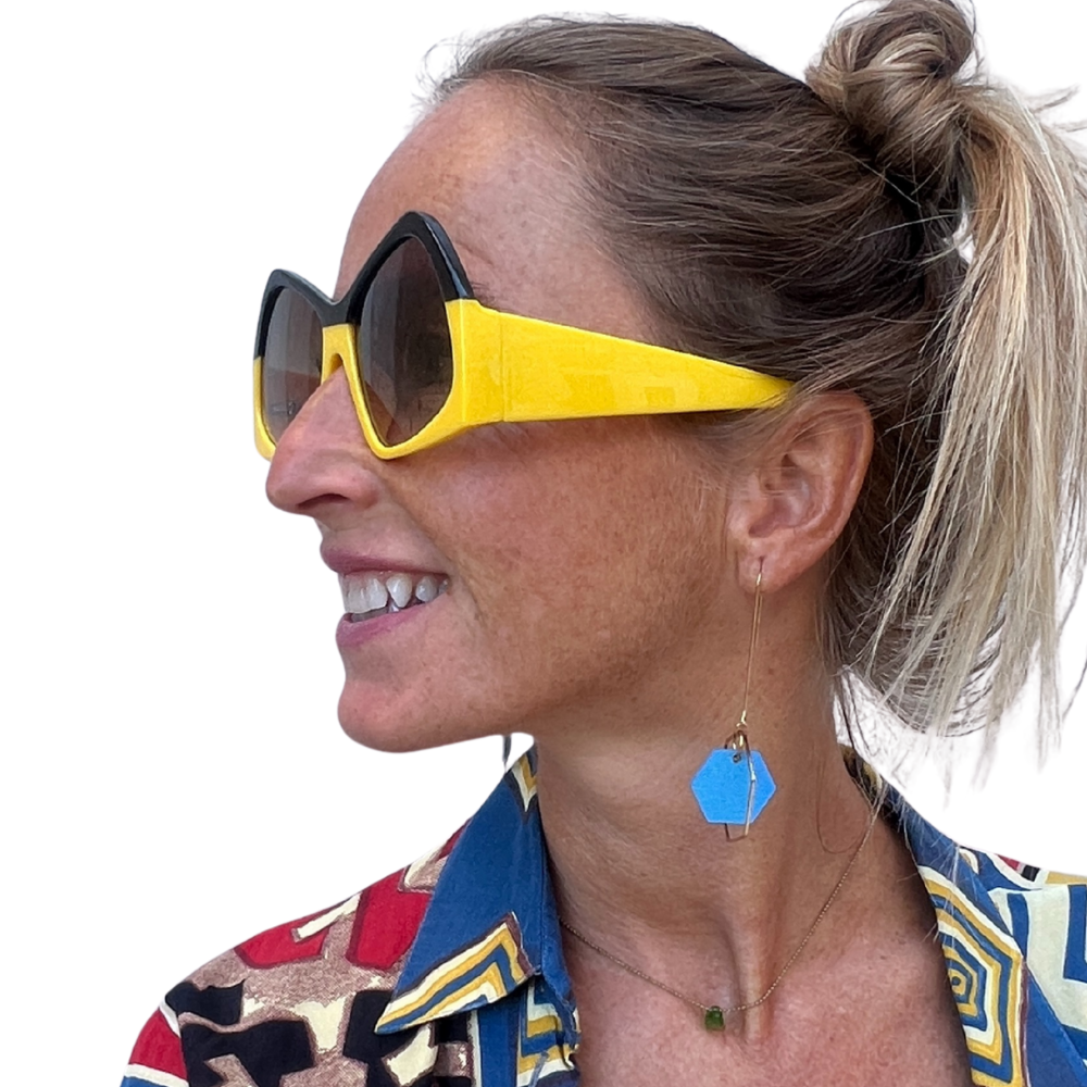 Woman wearing a colorful patterned shirt and yellow sunglasses with a blue earring.
