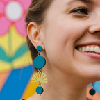 Woman wearing teal blue hand painted earrings with brass sunbursts standing in front of a flower mural