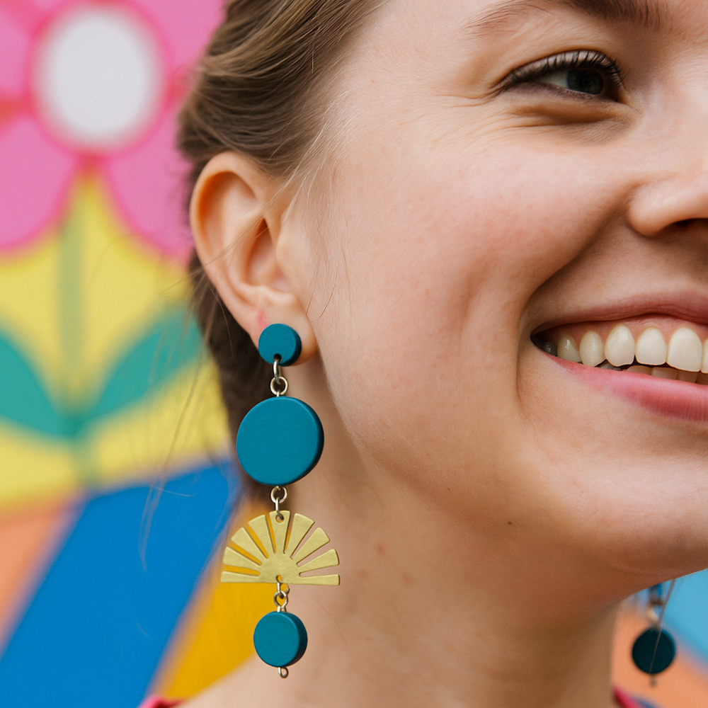 Woman wearing teal blue hand painted earrings with brass sunbursts standing in front of a flower mural